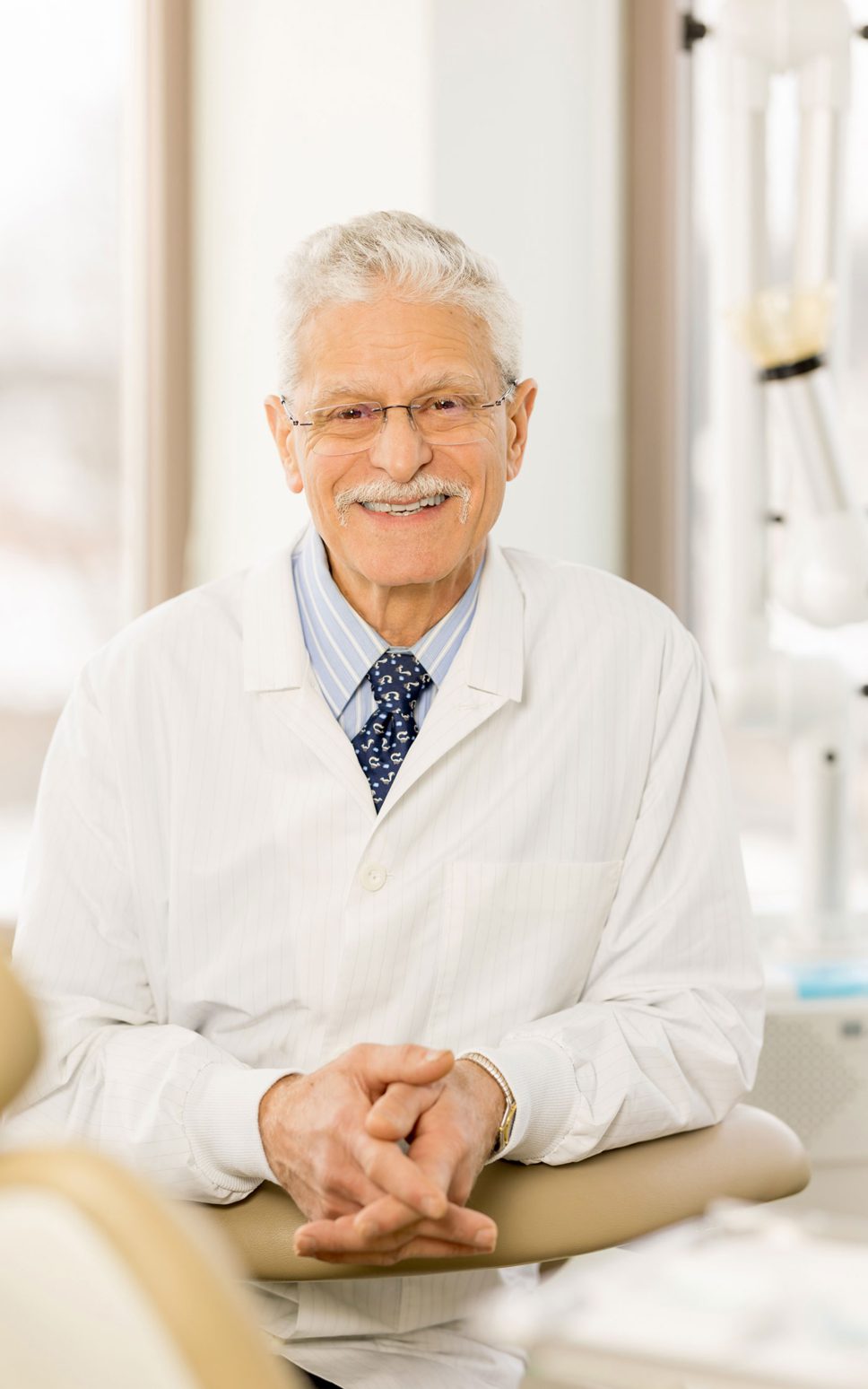 A man in a white lab coat and dark tie smiles with hands folded.