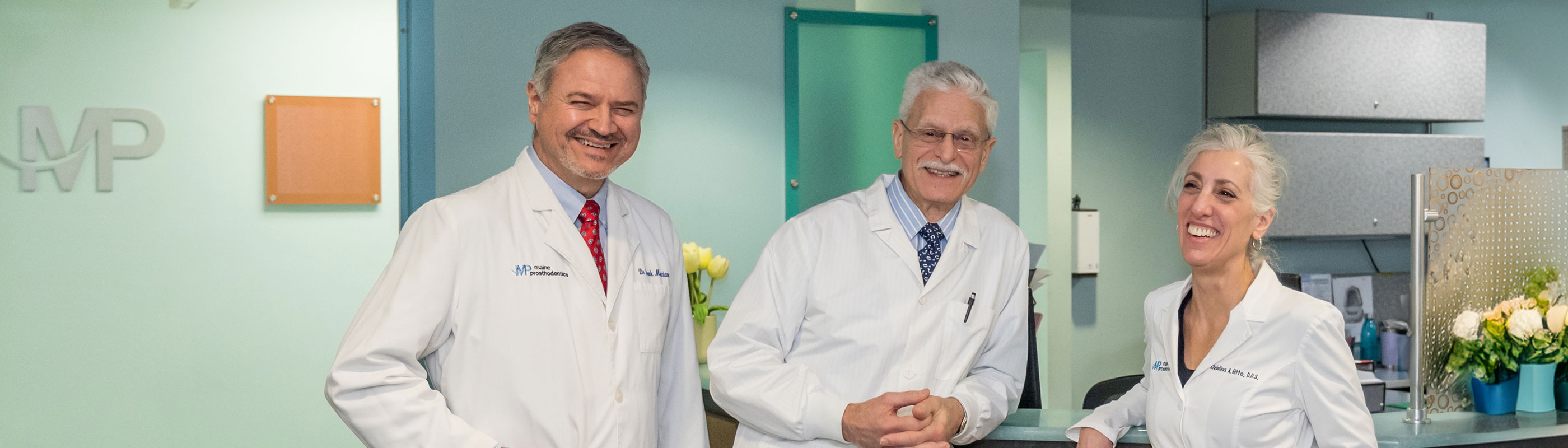 Three dentists in lab coats stand in front of a green and blue modern office reception desk.