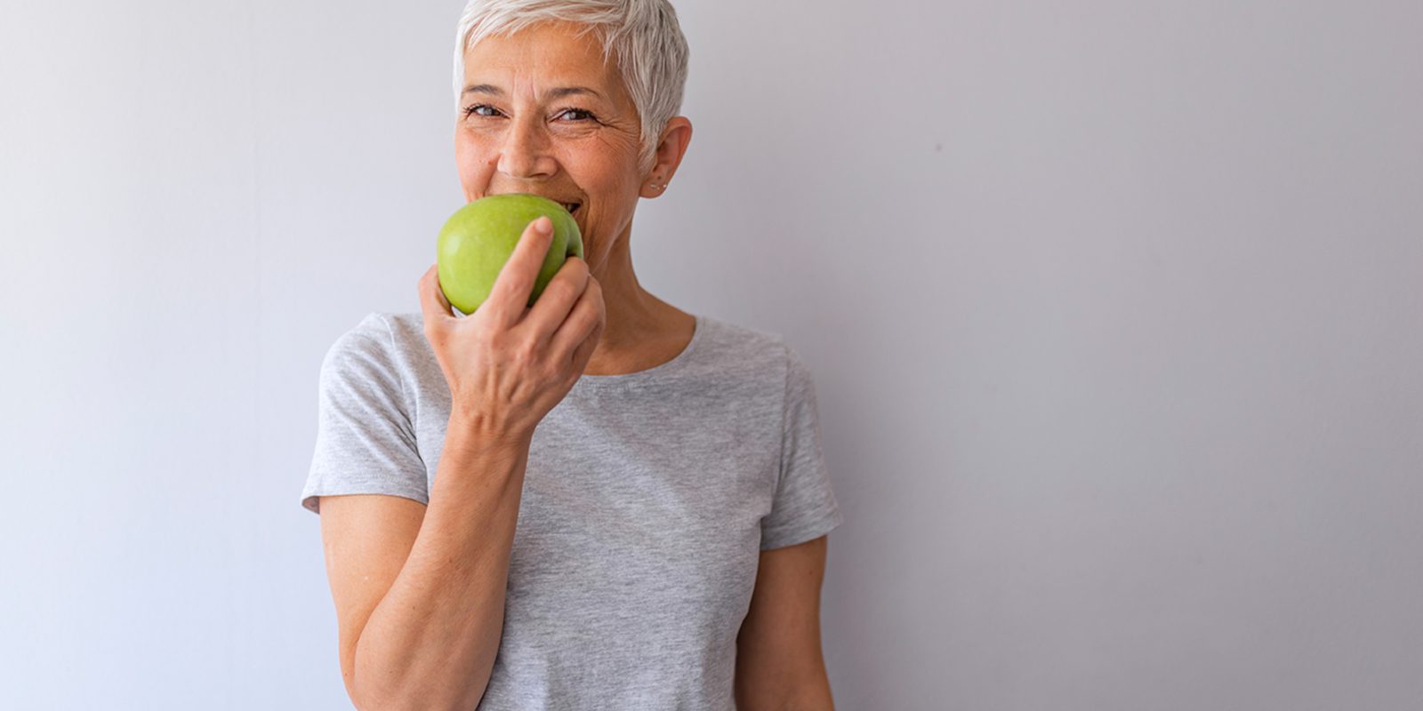 A woman with short gray hair smiles while biting into a bright green apple.