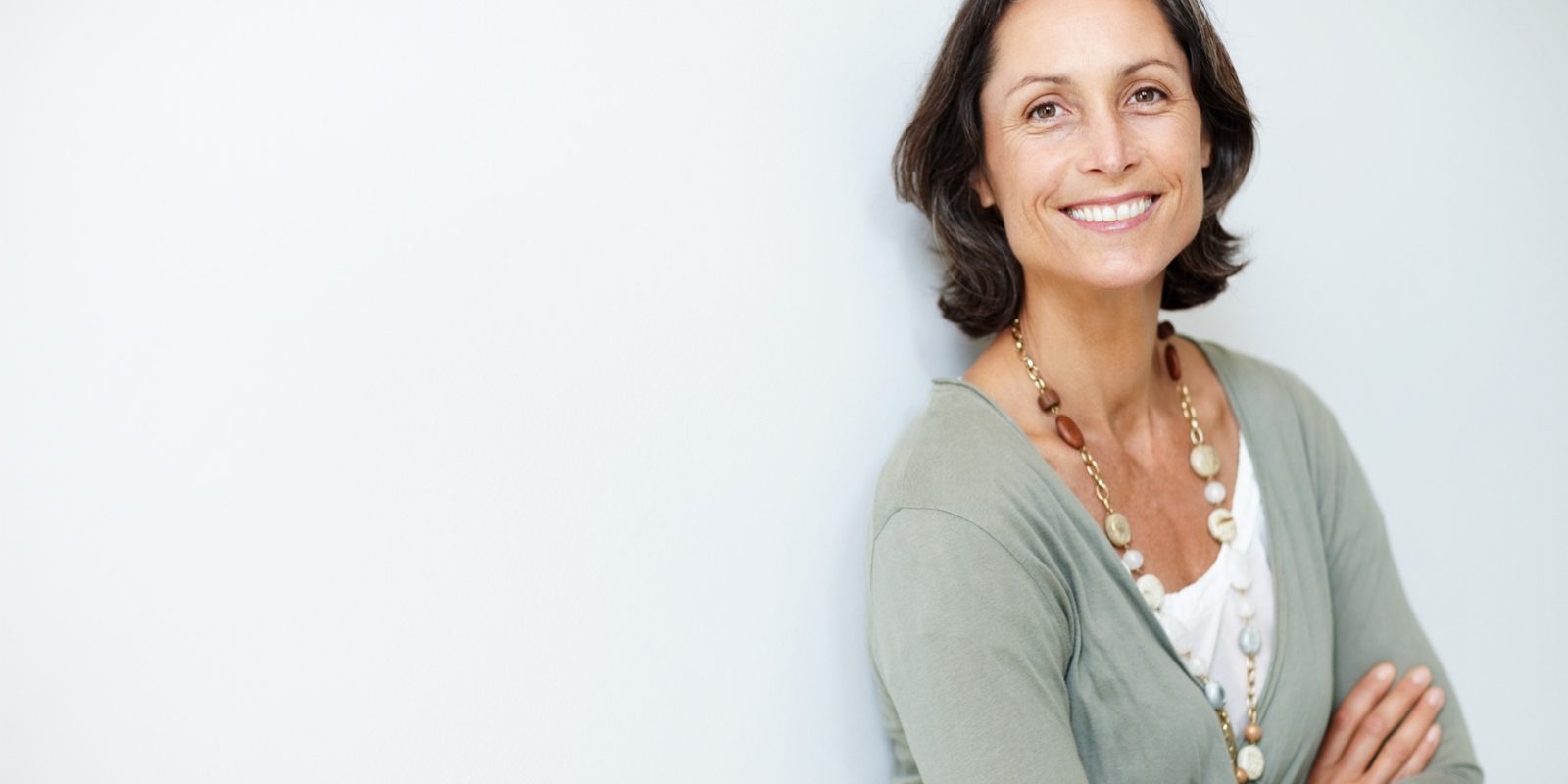 A woman in a soft green sweater leans against a white wall and smiles.