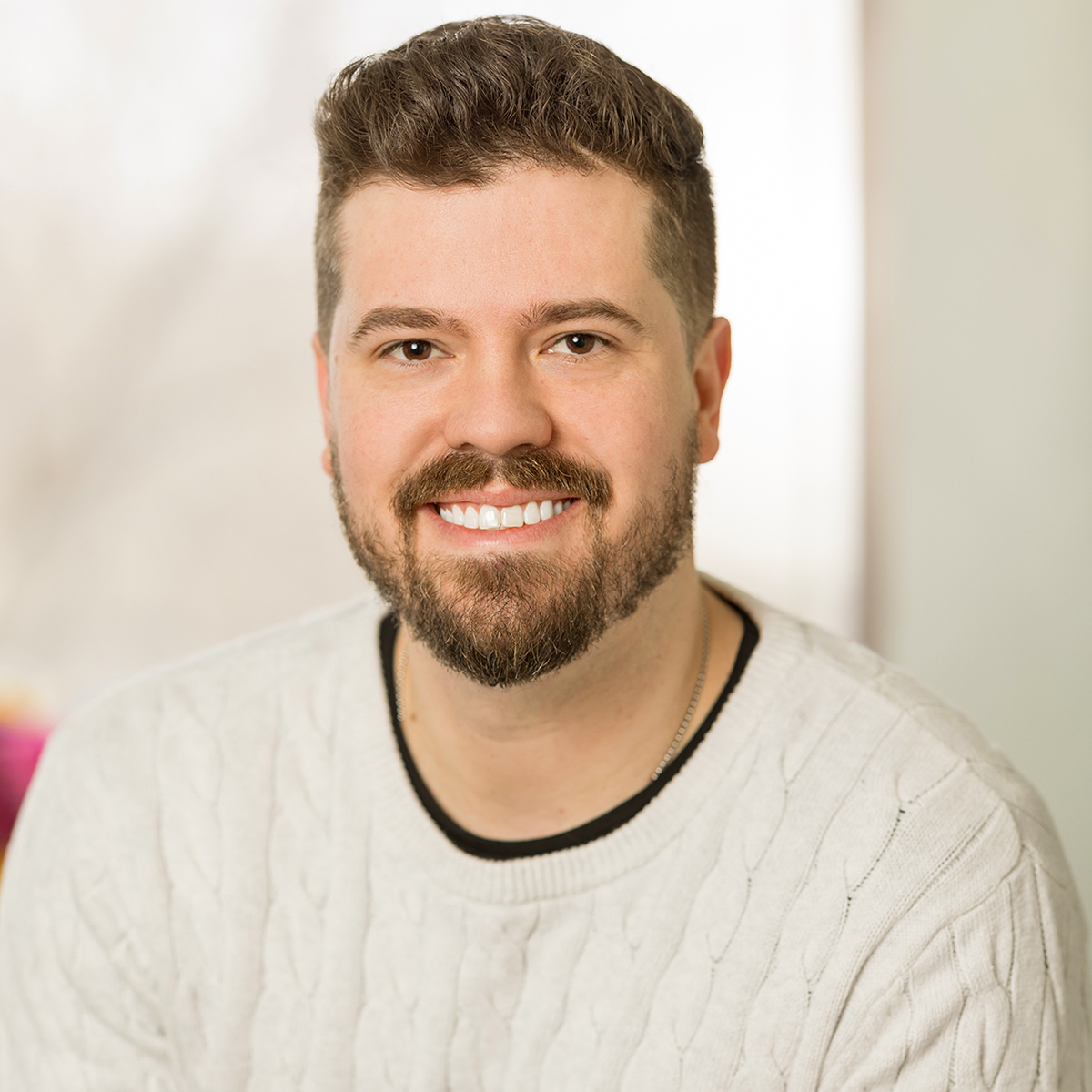 A bearded man in a white sweater smiles while leaning against his desk.